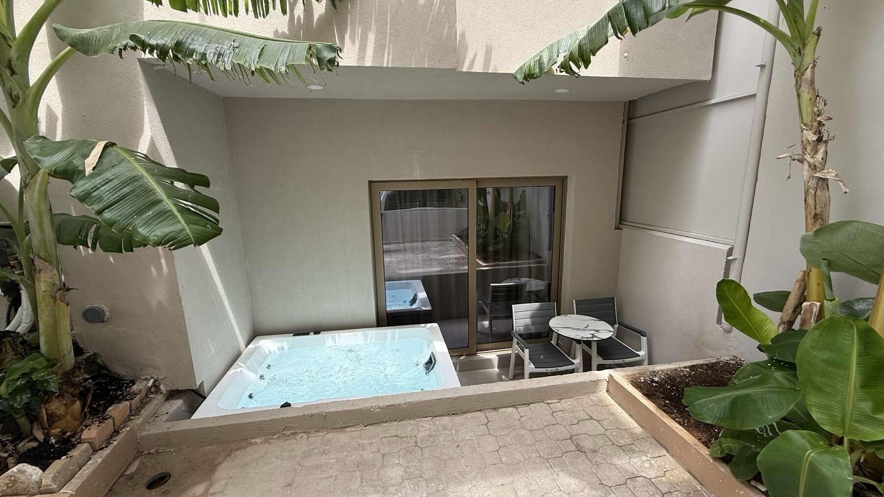 A small outdoor hot tub area with a glass door leading to a balcony, surrounded by large banana plants.