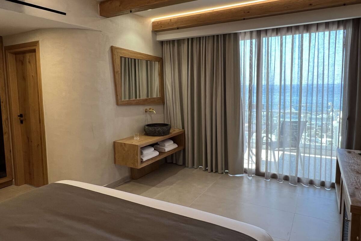 Une chambre d’hôtel moderne avec un grand lit, une coiffeuse en bois dotée d’un lavabo, un miroir et une vue sur la mer à travers de grandes rideaux.