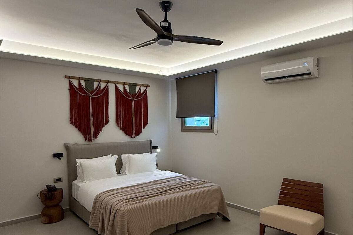 Une chambre à coucher comprenant un lit, un ventilateur de plafond, une fenêtre avec un store et un appareil de climatisation.