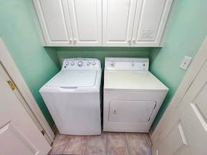 A laundry room with two white washing machines and a dryer, white cabinets above, and a green wall.