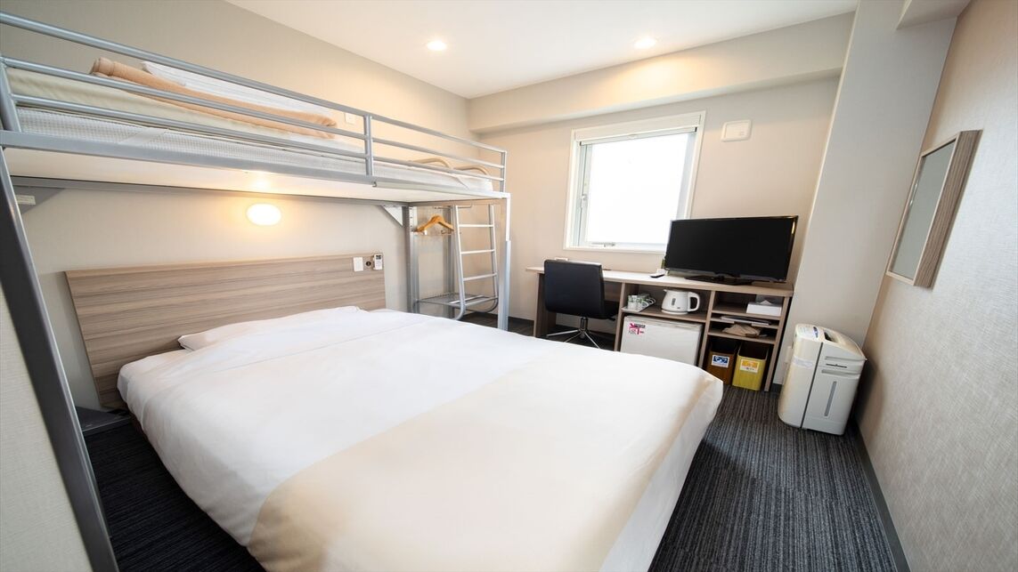 Een slaapkamer met een stapelbed, bureau, stoel, televisie en een klein keukenhoekje.