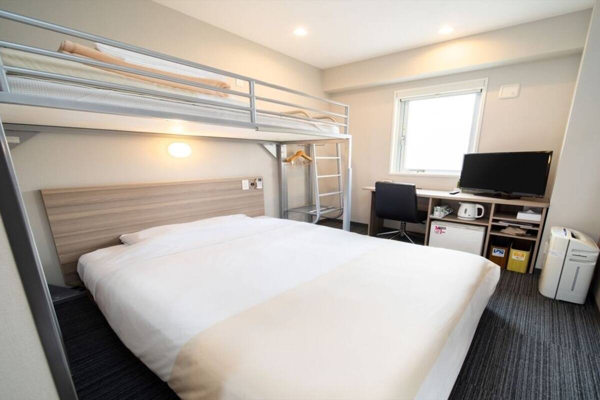 Une chambre avec un lit superposé, un bureau, une chaise, une télévision et une petite kitchenette.