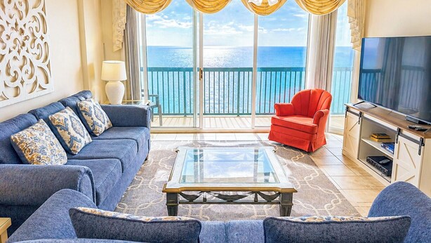 Una sala de estar con sofá, sillón y televisor, una mesita de centro de vidrio, y un balcón con vista al mar.