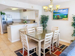 A dining area with a glass table and white chairs, a kitchen with white cabinets, and a painting of a landscape on the wall.