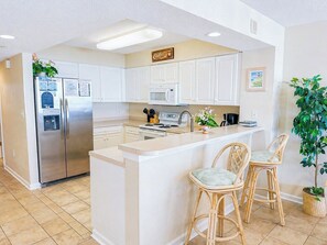 A kitchen with white cabinets, a stainless steel refrigerator, a microwave, a stove, and a bar area with two wicker chairs.