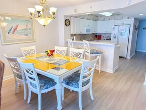 Un coin repas avec une table en verre et des chaises blanches, une cuisine avec des meubles blancs et, au fond, une porte bleue.