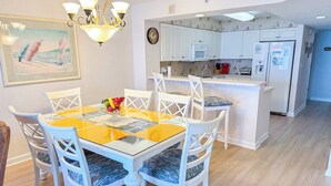 Un comedor con mesa de vidrio y sillas blancas, una cocina con muebles y electrodomésticos blancos, y un cuadro en la pared.