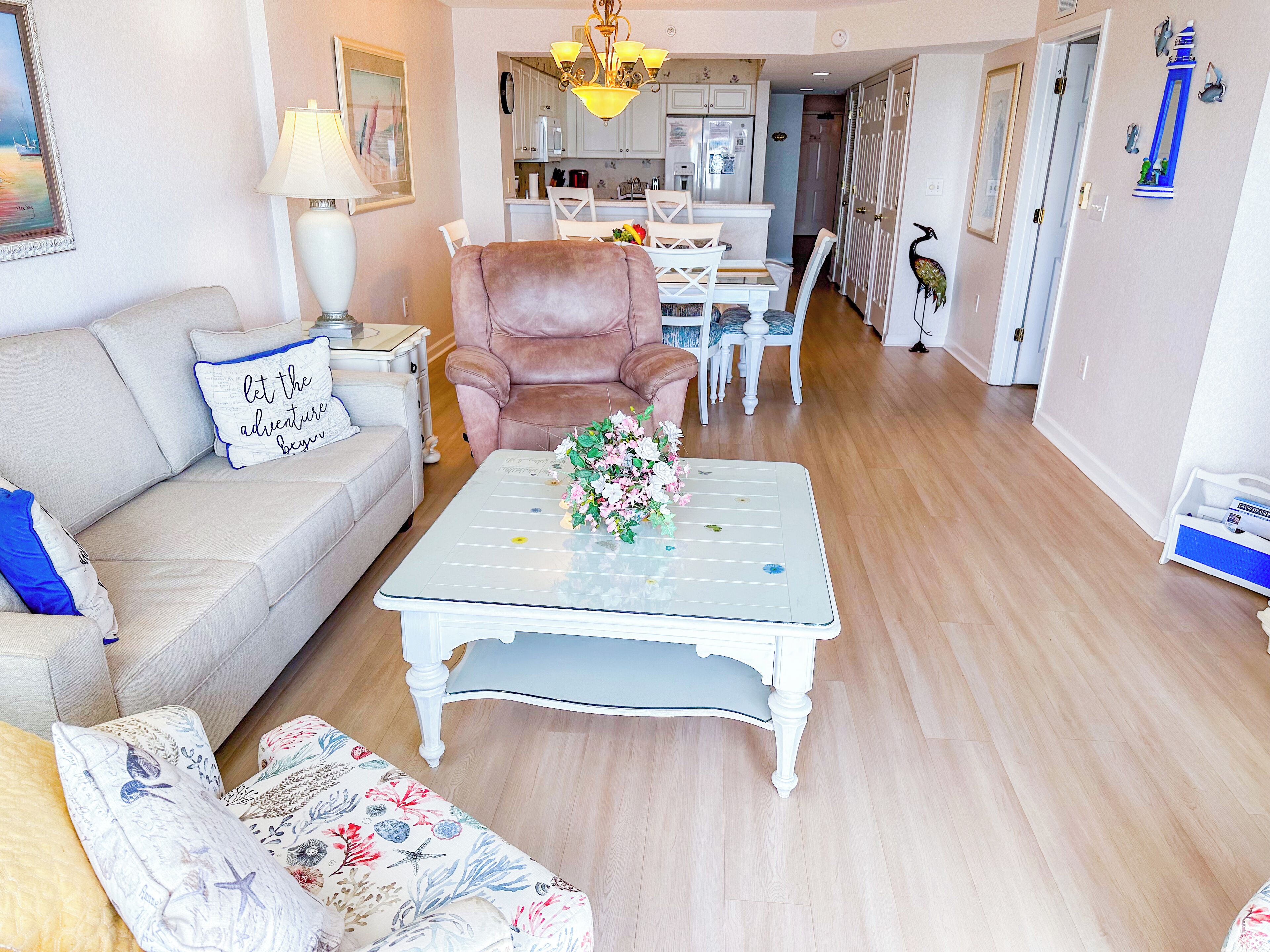 Een woonkamer met een bank, een fauteuil, een salontafel met bloemen, en een eethoek op de achtergrond.