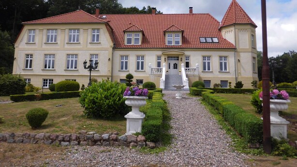 Ein großes, zweistöckiges Gebäude mit rotem Ziegeldach, zahlreichen Fenstern und einem zentralen Eingang mit Stufen.