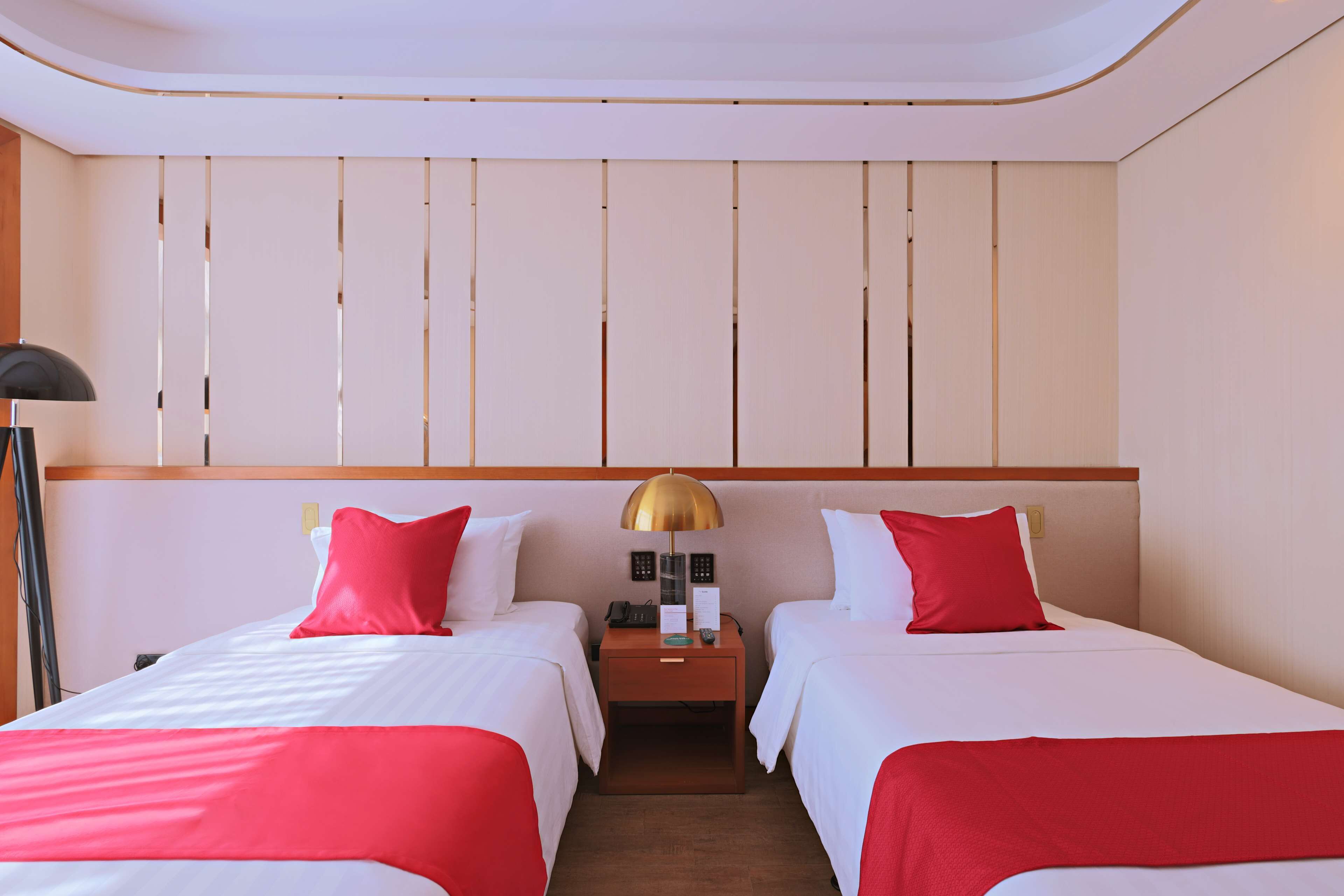 A hotel room with two beds, each with red and white bedding, a nightstand with a lamp, and a wooden headboard.