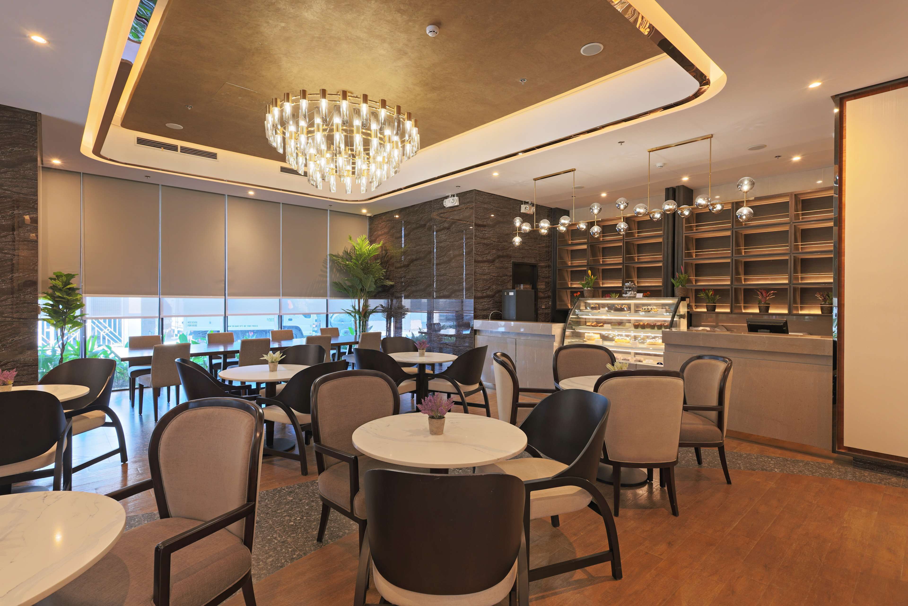 A modern restaurant with round tables and chairs, a central chandelier, and a counter with a display case.