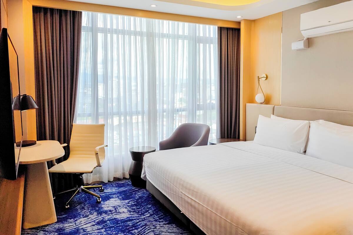 Ein Hotelzimmer mit einem großen Bett, einem Schreibtisch mit Stuhl, einem kleinen Tisch und einem großen Fenster mit Vorhängen.