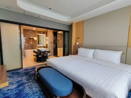 Habitación de hotel con una cama grande, un otoman azul redondo, un baño con lavabo y espejo, y un vestidor.