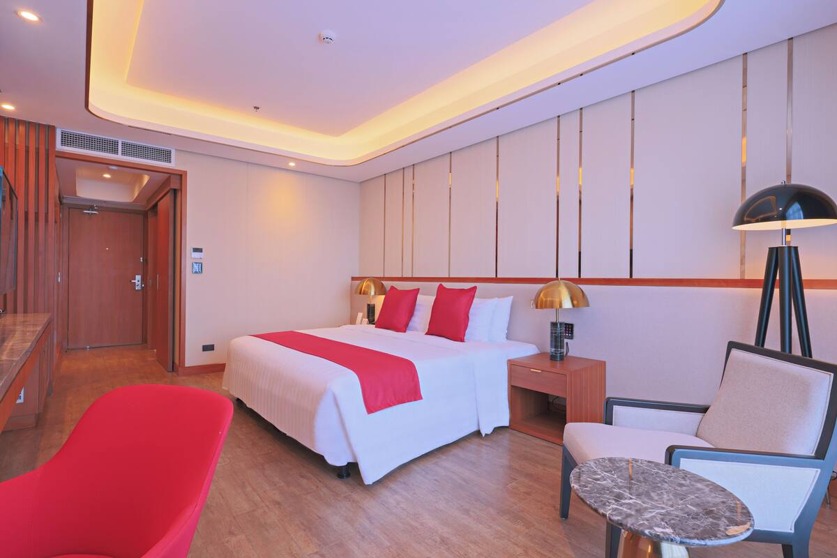Ein Hotelzimmer mit einem großen Bett, einem roten Sessel, einem kleinen Tisch und einem Nachttisch mit Lampe.