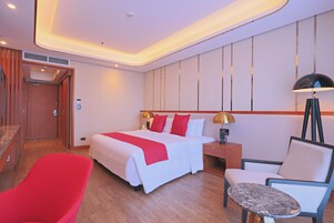 Una habitación de hotel con una cama grande, una silla roja, una mesita pequeña y una mesita de noche con una lámpara.