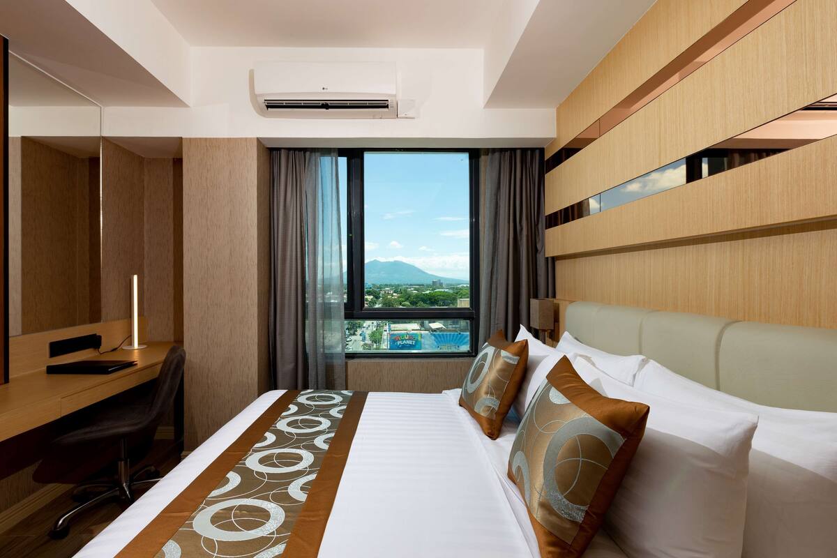 Ein Hotelzimmer mit Bett, Schreibtisch, Stuhl, großem Fenster und Klimaanlage, mit Blick auf Berge.