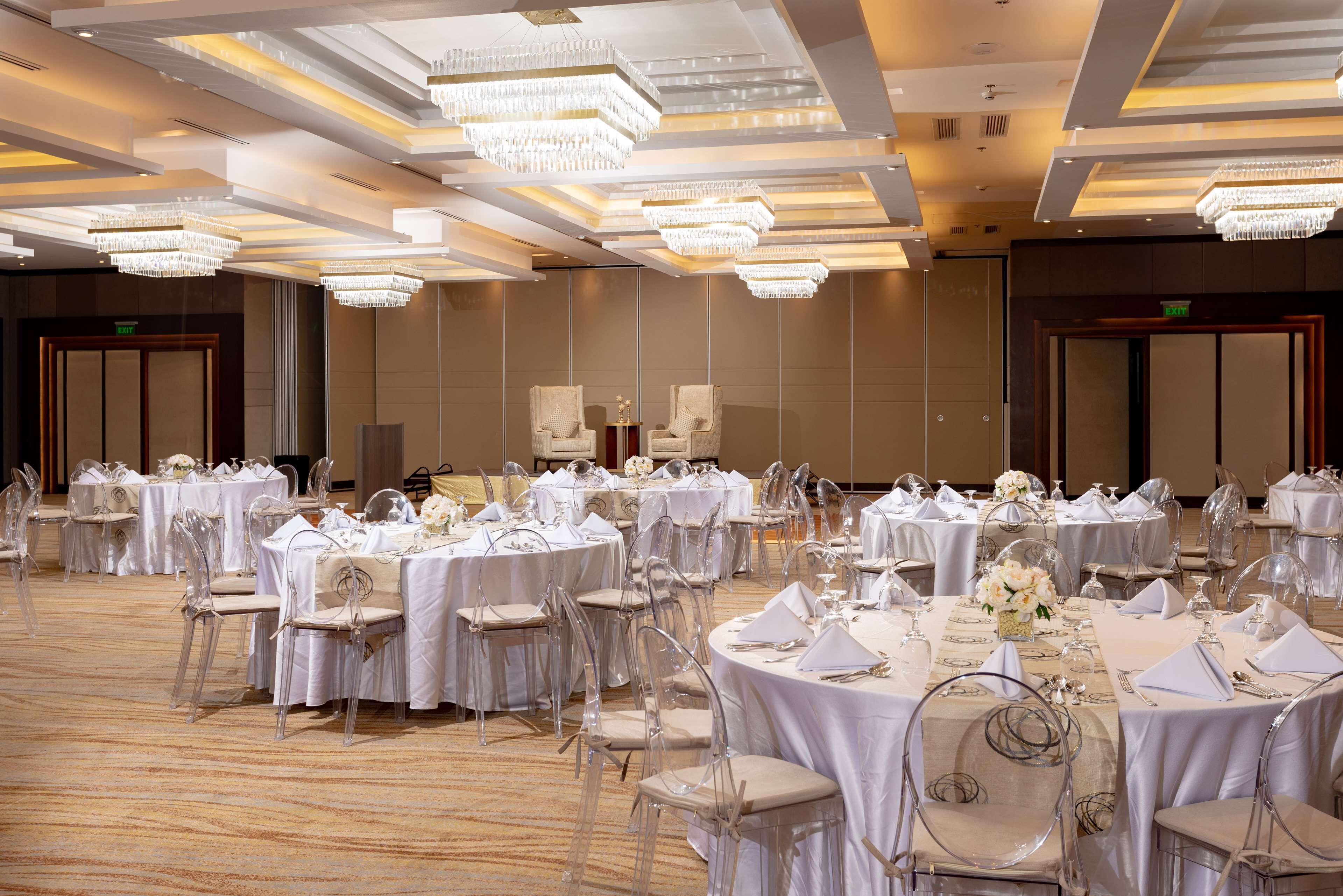 Un salón de banquetes con mesas redondas dispuestas para eventos, sillas transparentes y candelabros.