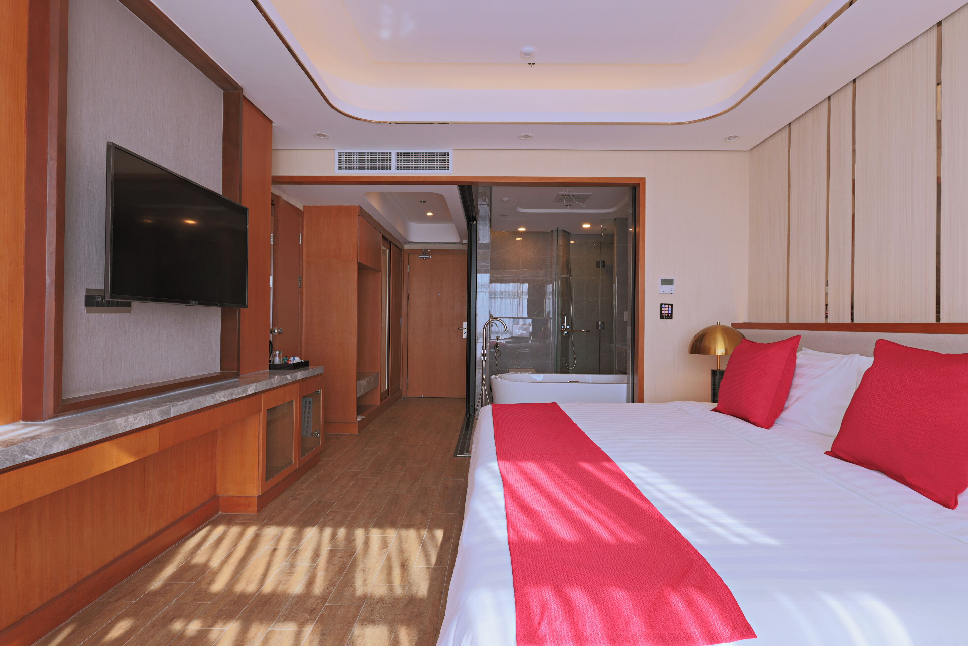A hotel room with a large bed, a TV mounted on the wall, a bathroom with a glass shower, and a wooden cabinet.