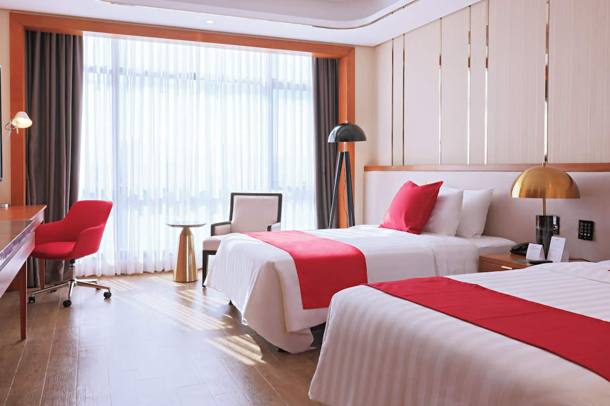 Ein Hotelzimmer mit zwei Betten, einem Schreibtisch, einem Stuhl und Blick auf die Stadt durch große Fenster.