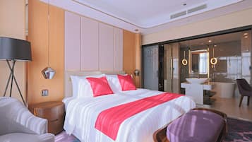 A hotel room with a large bed, a bedside table, a chair, a desk with a chair, and a bathroom visible through a glass door.