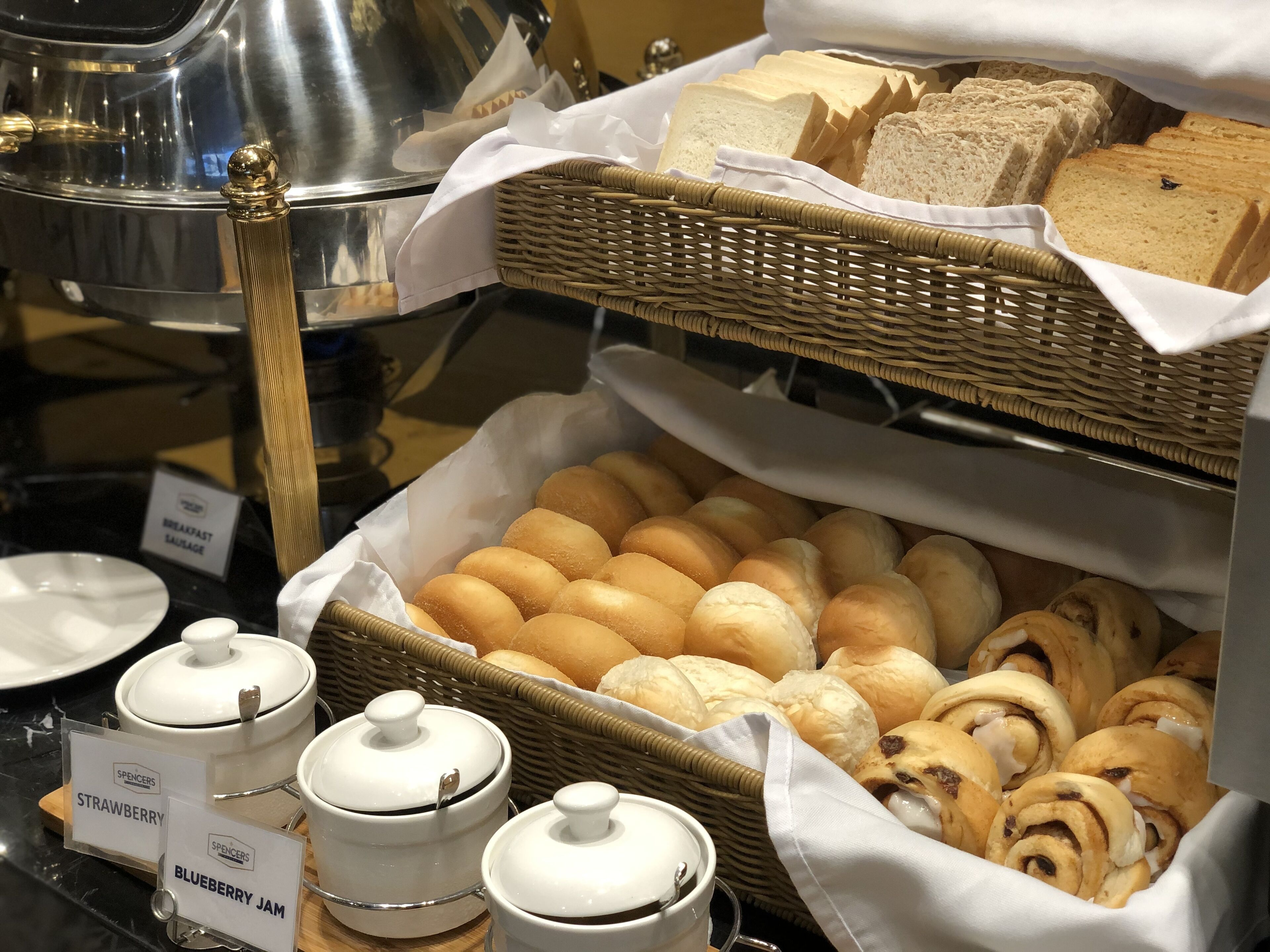 Een ontbijtbuffet met diverse broden en gebak, waaronder broodjes en kaneelbroodjes, plus verpakkingen met aardbeien- en bosbessenjam.