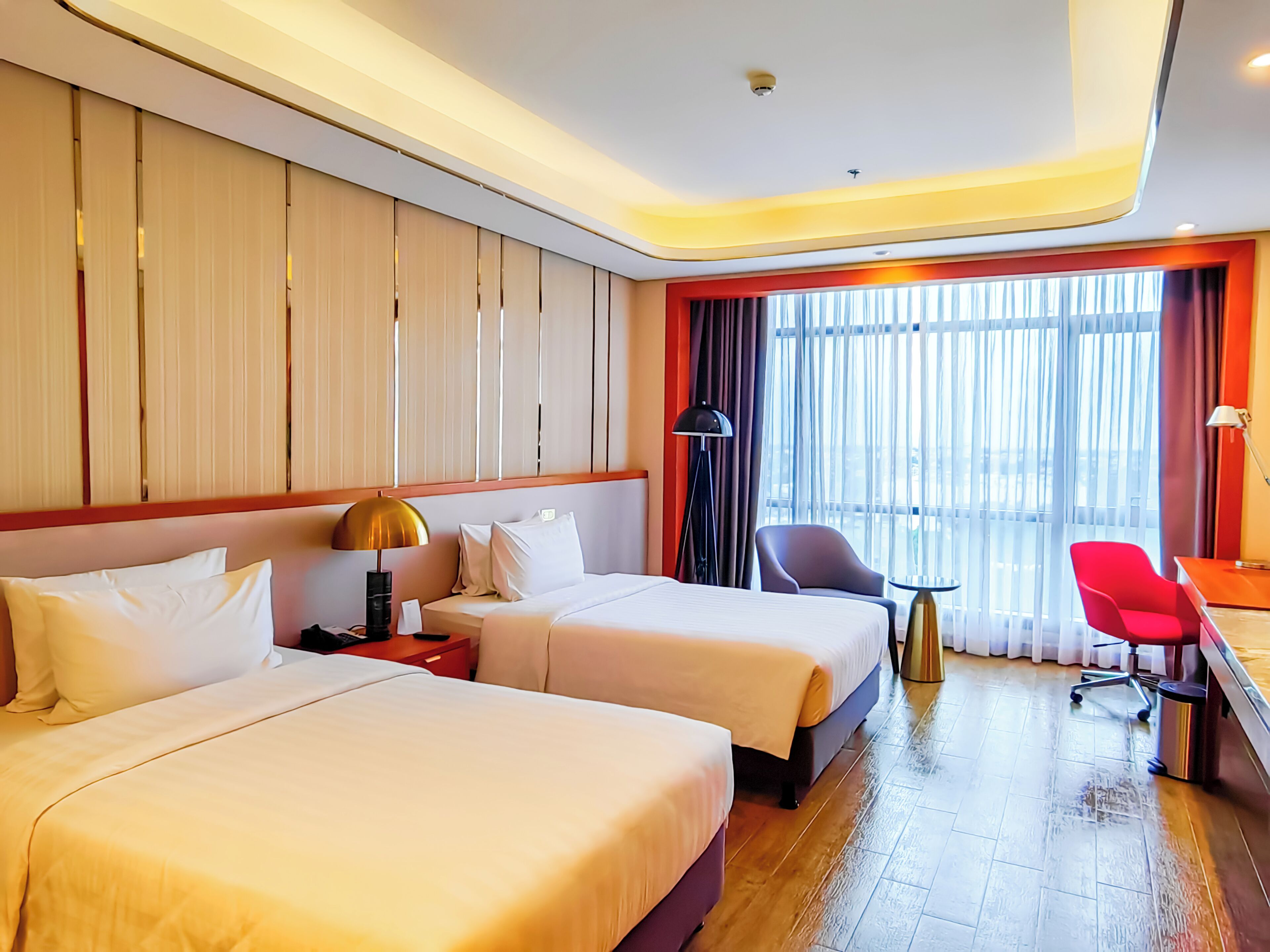 Een hotelkamer met twee bedden, een bureau, een stoel en uitzicht op de stad door grote ramen.