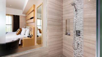 A modern hotel room with a large bed, a sitting area, a shower with a glass enclosure, and a large mirror reflecting the room.