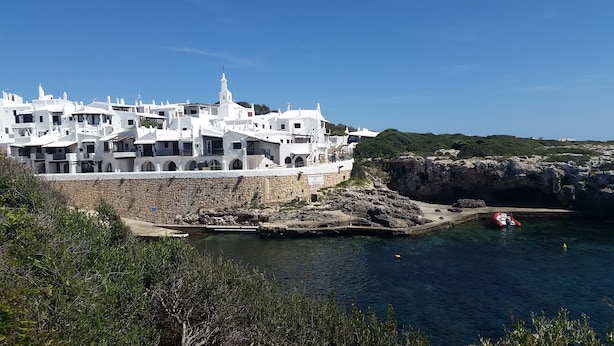 A coastal building with white walls and a stone base, situated near a rocky shoreline with clear blue water. - Comfortable Villa in Binibeca with Private Pool and Roof Terrace with Sea Views (Menorca)