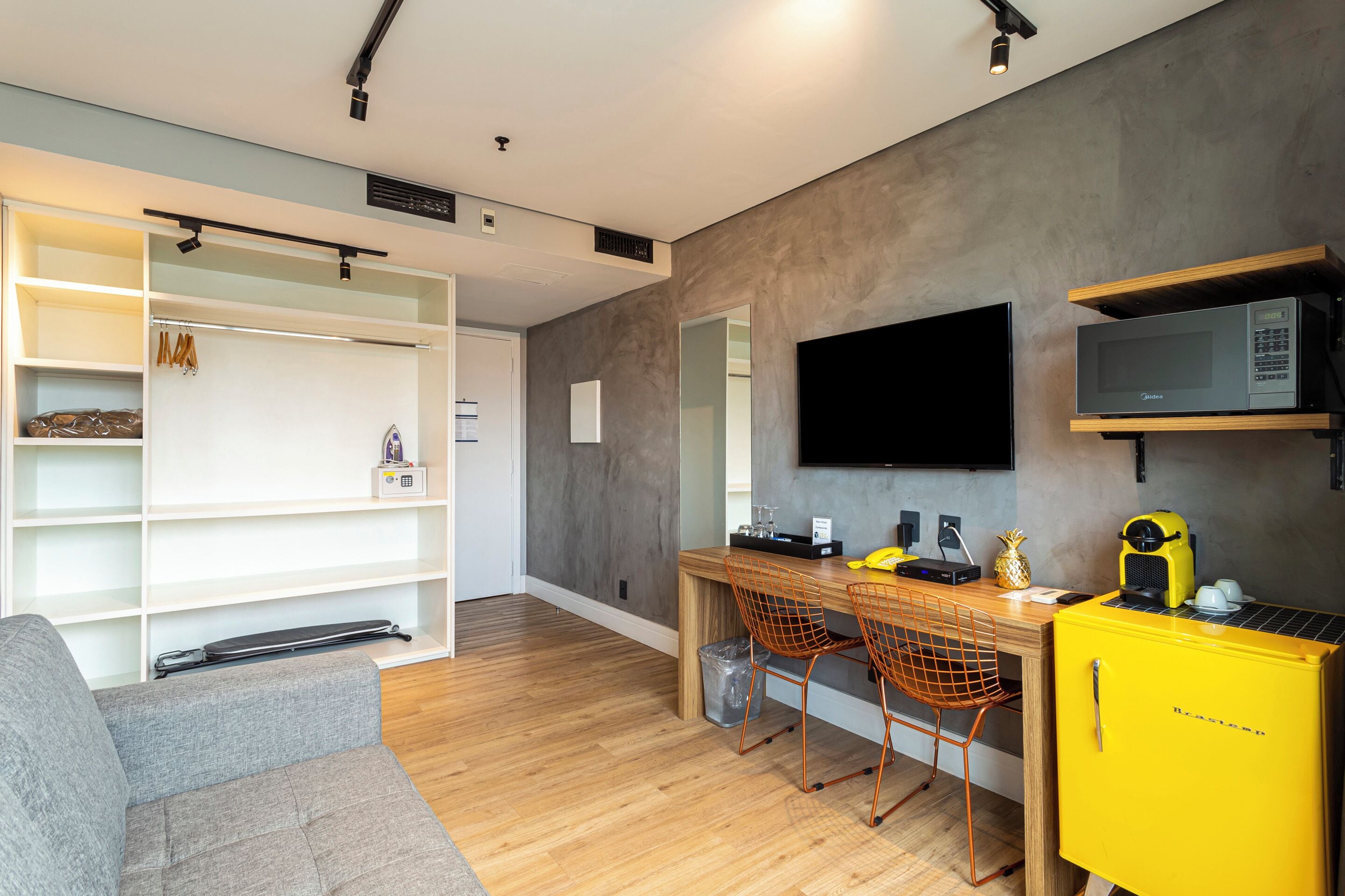A modern living room with a grey sofa, wooden flooring, a dining area with a yellow refrigerator, and a flat-screen TV mounted on the wall.