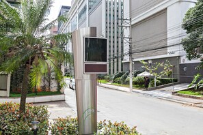 Ein modernes Gebäude mit großer Glasfassade, einem Metallpfosten mit digitaler Anzeige und einem gepflegten Garten mit Palmen und Sträuchern.