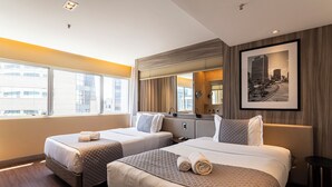 Ein Hotelzimmer mit zwei Betten, einem großen Fenster, Blick auf die Stadt und einem gerahmten Bild an der Wand.