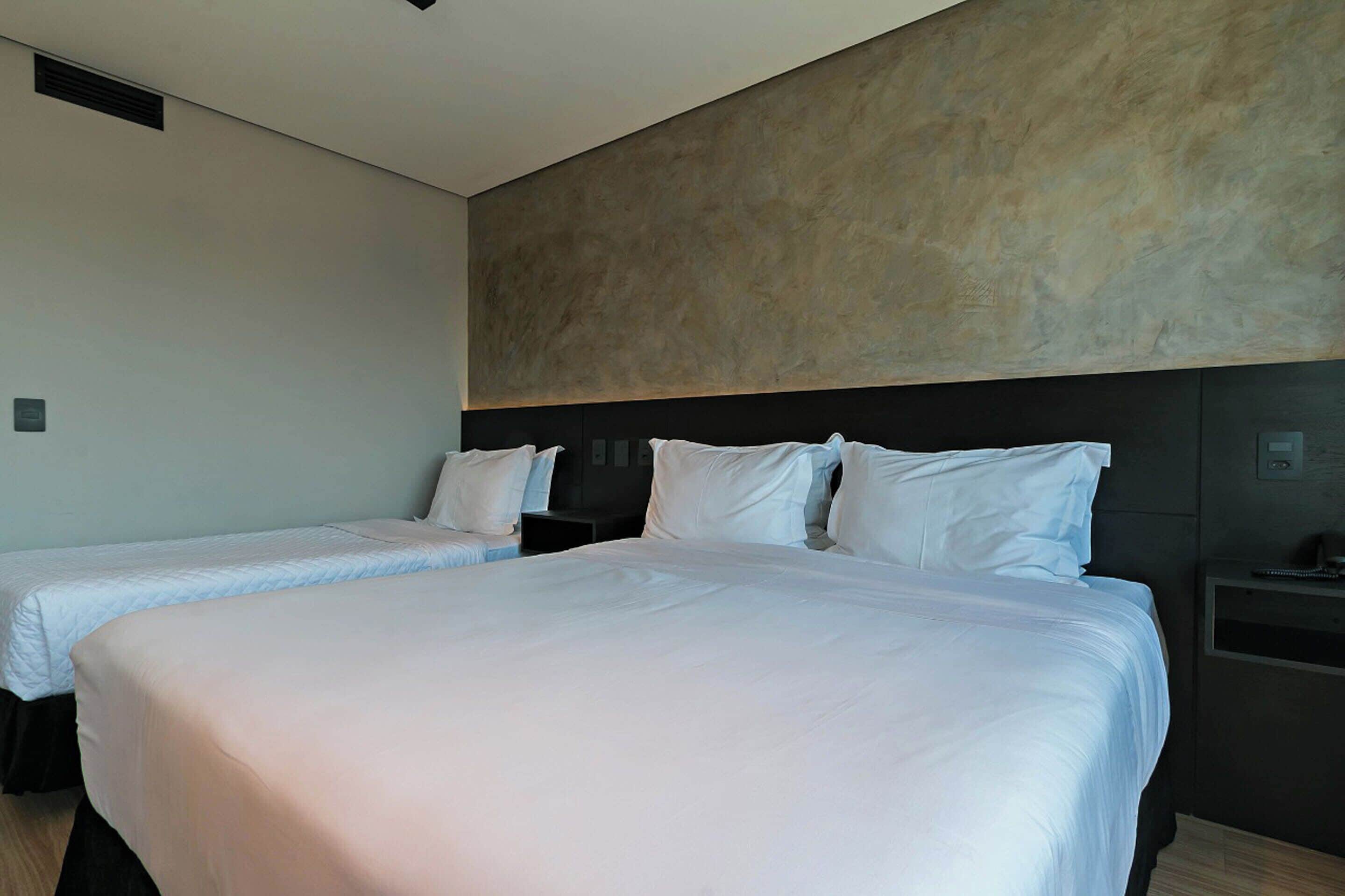 A hotel room with two beds, a large wall-mounted headboard, and a small bedside table with a lamp.