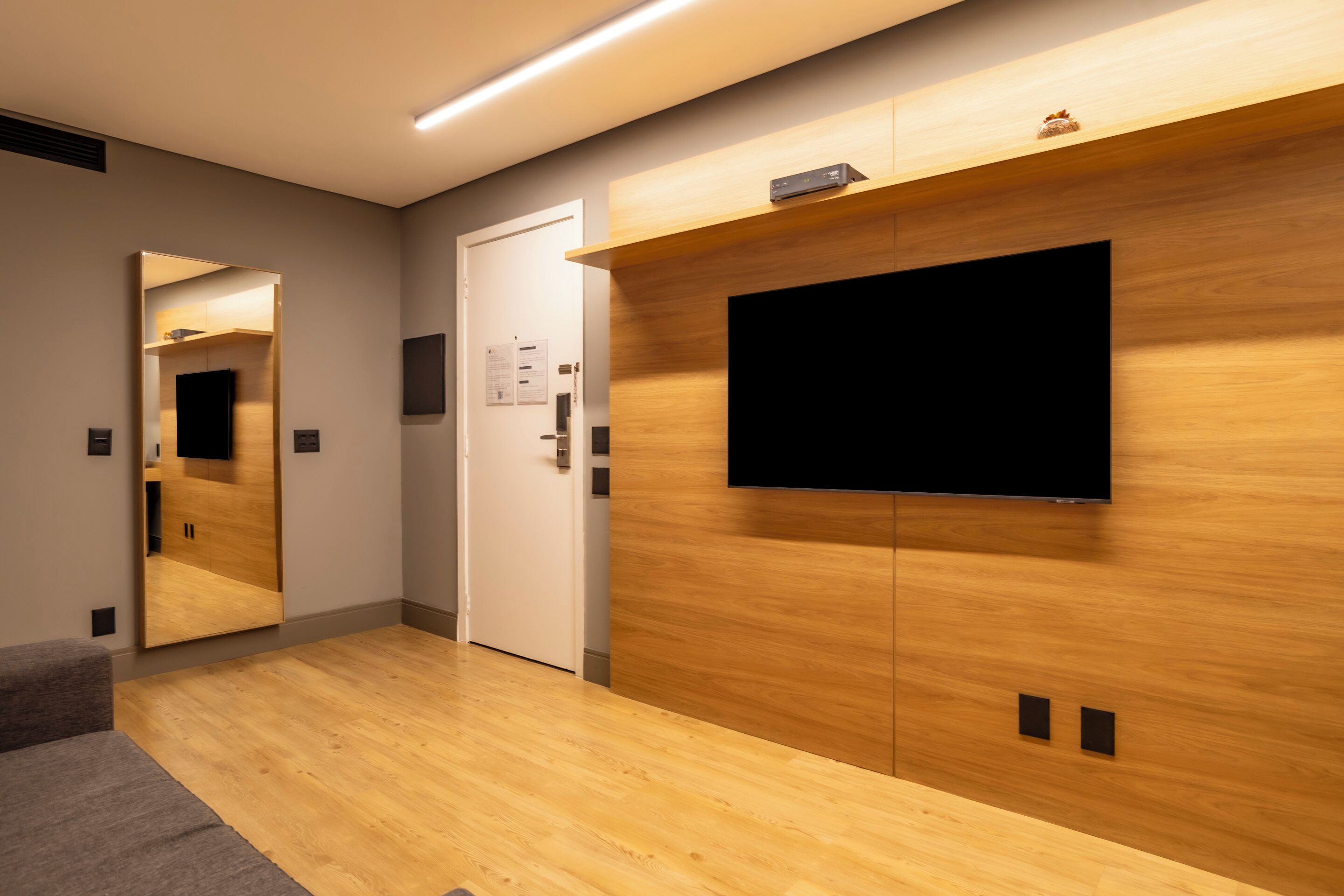 Una habitación de hotel moderna con piso de madera, un espejo grande, un televisor de pantalla plana montado en la pared y una puerta con picaporte.