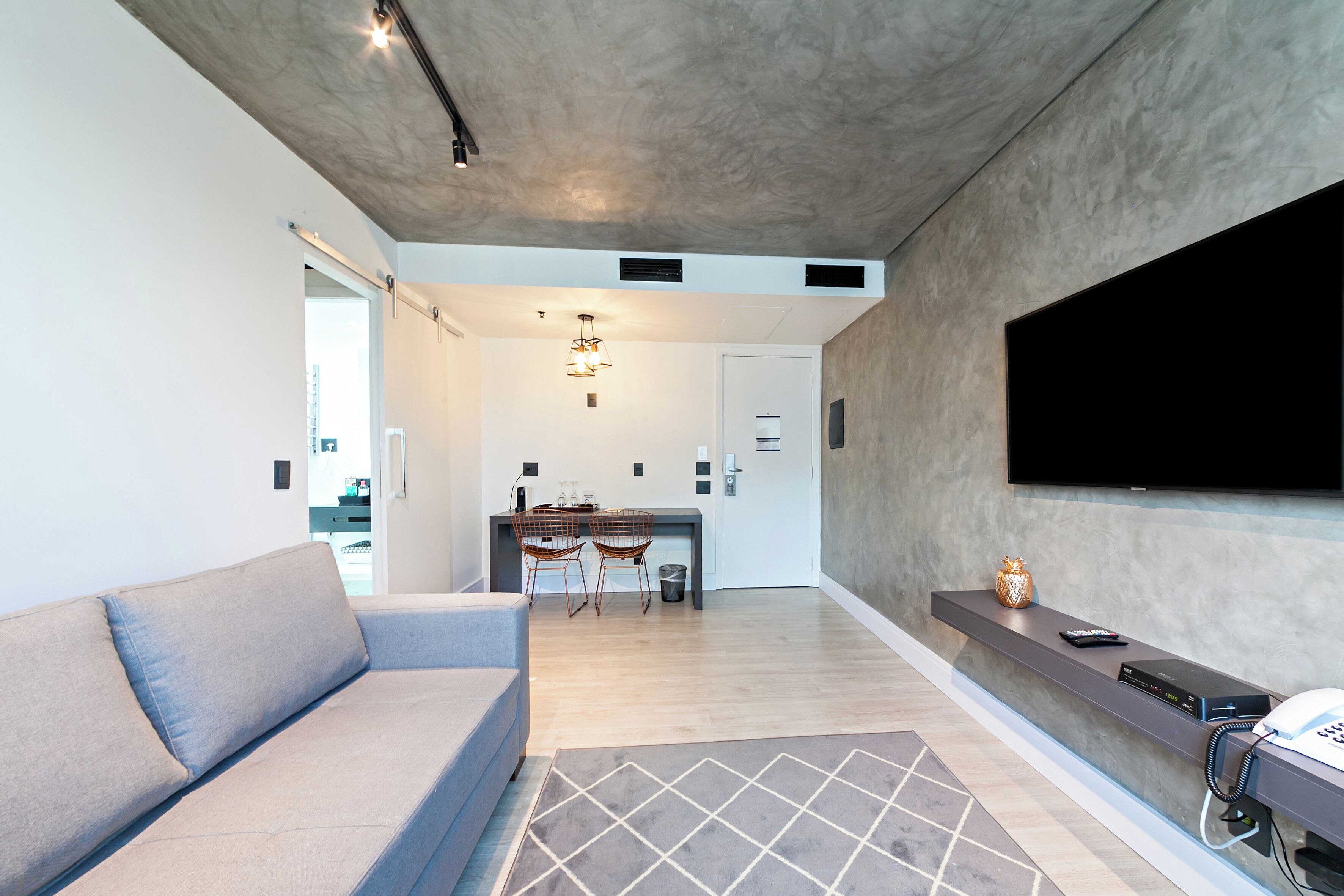 A modern living room with a grey sofa, a dining area with a table and chairs, a flat-screen TV mounted on the wall, and a built-in phone station.