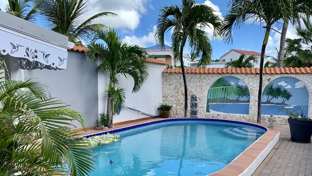 A swimming pool with a curved edge, surrounded by palm trees and a stone wall with a mural.