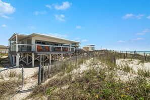 Una propiedad frente al mar con una casa moderna sobre pilotes, una terraza de madera y una playa arenosa al fondo.