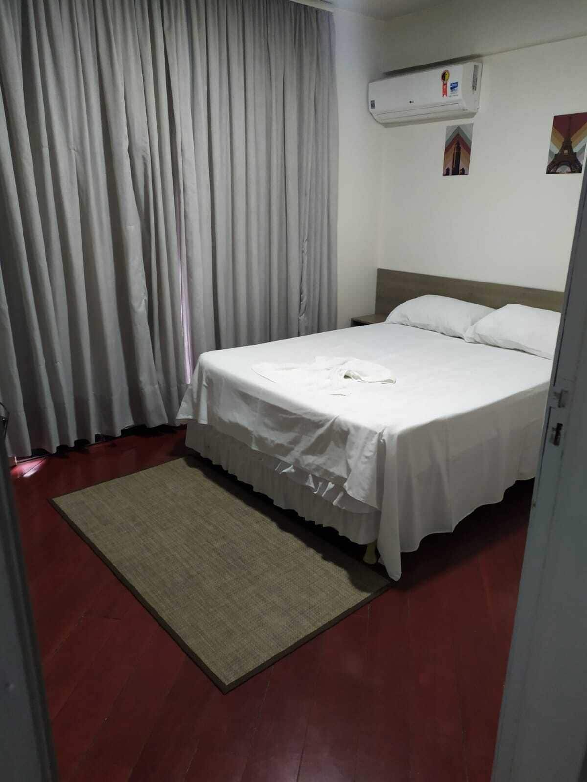 A hotel room with a bed, a white curtain, an air conditioning unit, and a small rug.