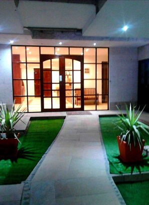 A modern building entrance with a glass door, a tiled pathway, and green artificial grass on either side.