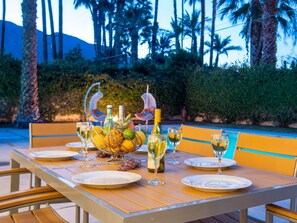 Une table de jardin, dressée pour un repas en plein air, avec des assiettes, des verres, un saladier rempli de fruits et des bouteilles.