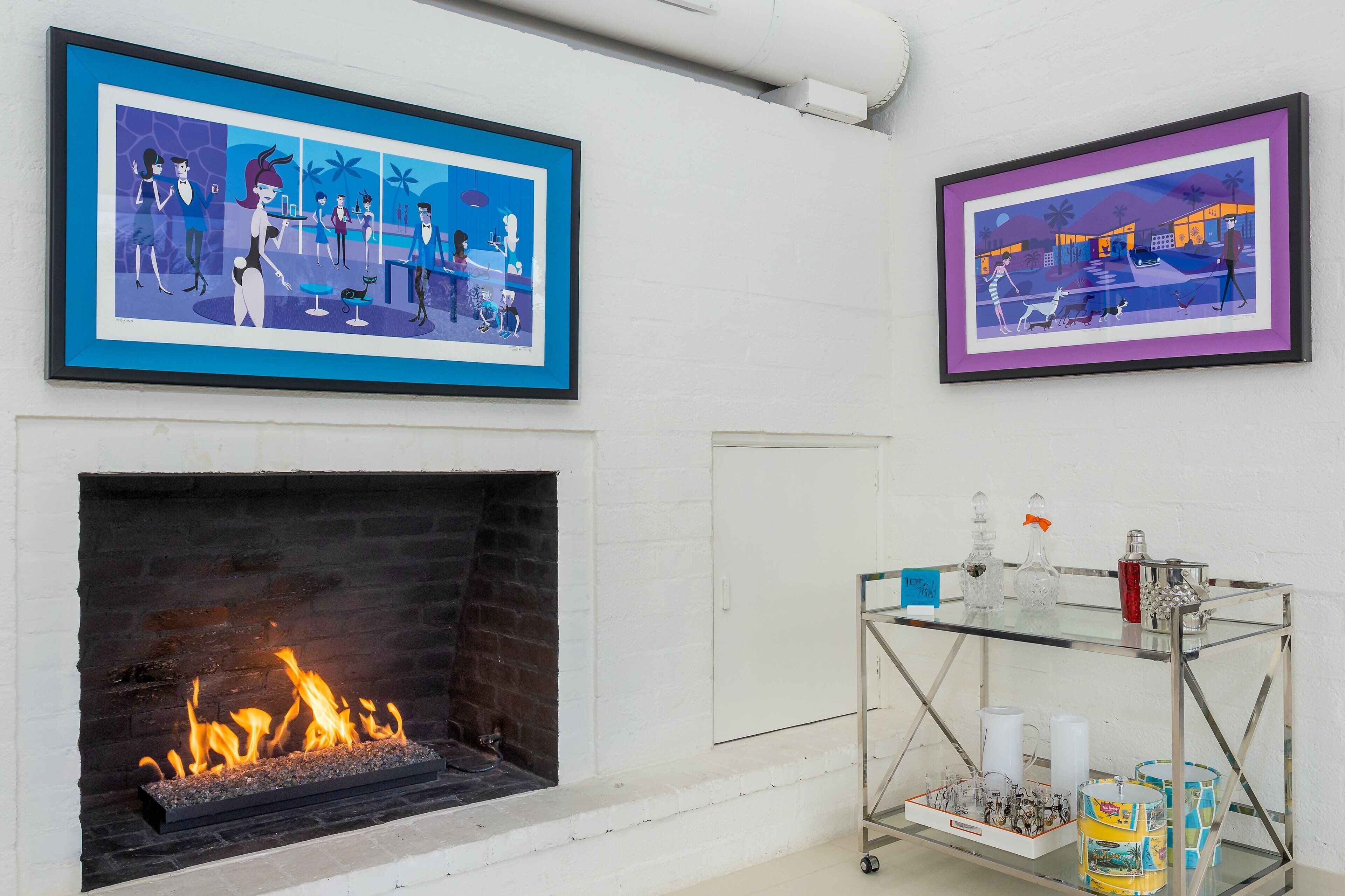 A room with a fireplace, two framed artworks, and a glass display case with bottles.
