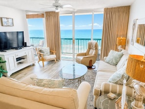 A spacious living room with a large flat-screen TV, comfortable seating, a glass coffee table, and a view of the ocean through large windows.