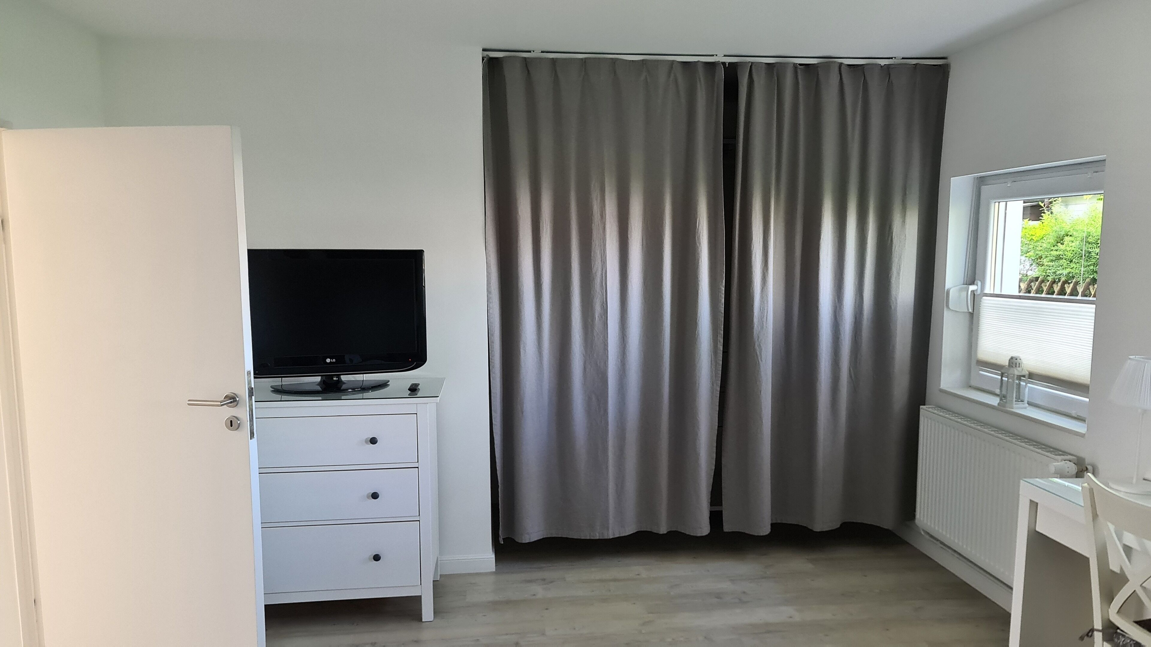 Ein Zimmer mit einem weißen Schrank, einem Flachbildfernseher darauf, einem grauen Vorhang und einem Fenster mit Blick auf Grünflächen.