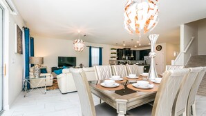 A dining area with a large table set for six, surrounded by six chairs. There is a kitchen in the background with modern appliances. - Champions Gate Exec Villas- 9085hs (Davenport)