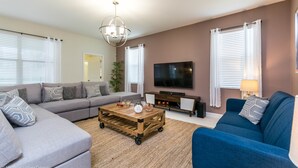 A modern living room with a sectional sofa, a blue armchair, a wooden coffee table, a flat-screen TV, and a chandelier. - Champions Gate Exec Villas- 9026sd (Davenport)