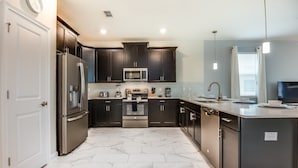 A modern kitchen with dark cabinetry, stainless steel appliances, and a marble floor. - Champions Gate Exec Villas- 1586sandb (Davenport)
