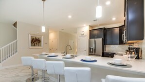 A modern kitchen with dark cabinets, a white countertop, and a bar area with white stools. - Champions Gate Exec Villas- 1586sandb (Davenport)