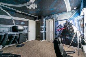 Quarto com mural temático do espaço, uma escrivaninha com computador e uma cadeira.