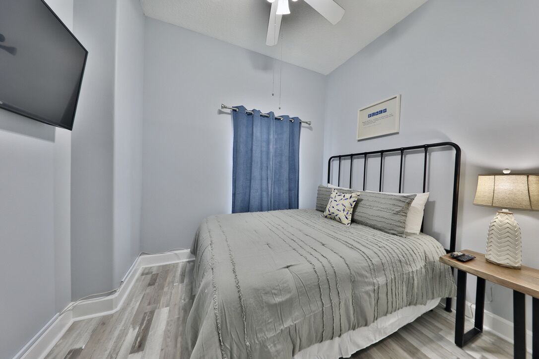 Un dormitorio con cama, mesita de noche, lámpara, ventilador de techo y televisor montado en la pared.