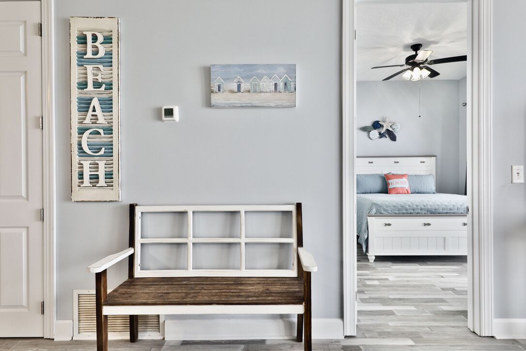 Una habitación con un banco de madera, un letrero en la pared que dice "BEACH" y una cama con cabecera blanca y ventilador de techo.