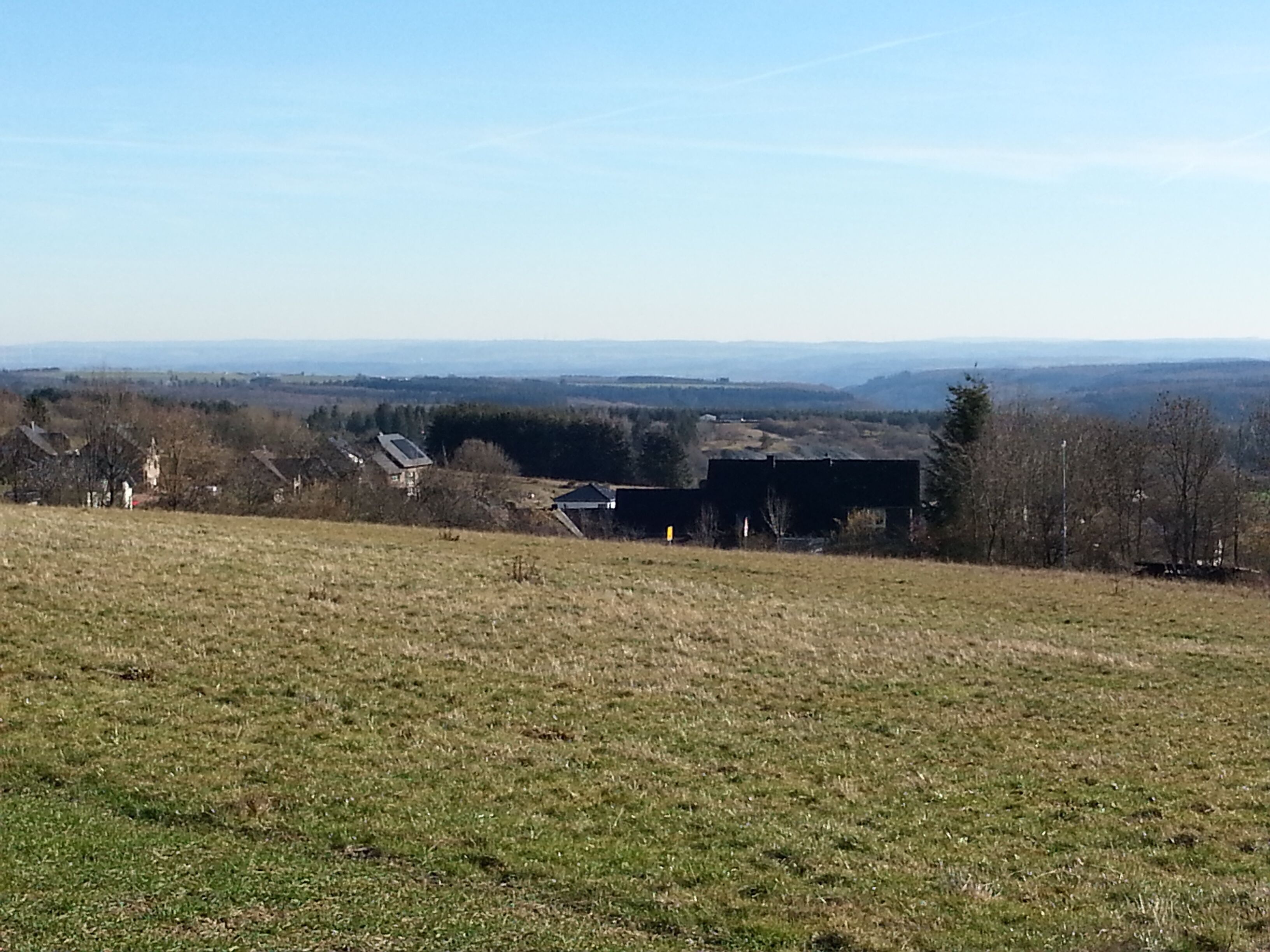 A rural landscape with a grassy field, a few buildings, and a distant view of hills.